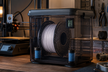Nylon filament spool stored in a sealed dry box with desiccant packs and a humidity monitor beside a 3D printer.