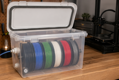 Clear plastic bin converted into an airtight filament container holding multiple spools with a sealed lid and feed-through fittings in a 3D printing workspace